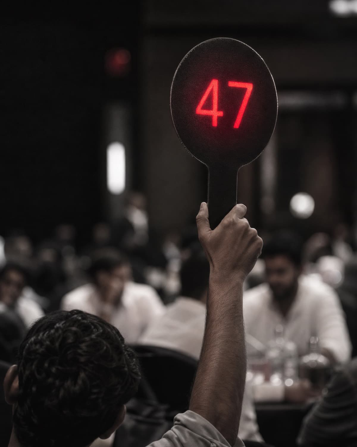 A hand raising a numbered bidding paddle glowing red above a dim auction hall